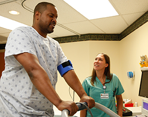 Proveedor de atención médica realizando una prueba de esfuerzo cardíaco a un hombre sobre una cinta caminadora en la sala de examinación.