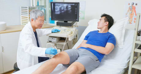 Proveedor de atención médica colocando transductor sobre la rodilla de una persona, con imágenes que se muestran en el monitor de una computadora.